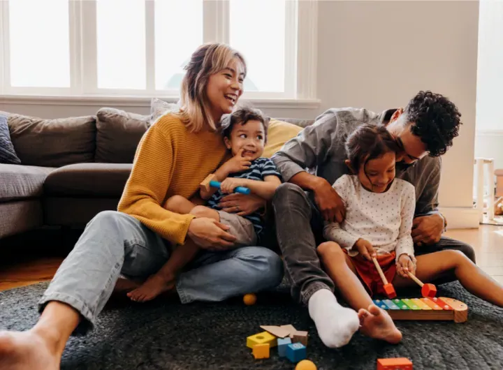 A young family sits on the floor, laughing and playing together with toys in a cosy living room.