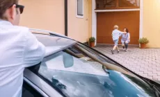 A father watches from his car as his young son runs to hug his mother in front of their home.